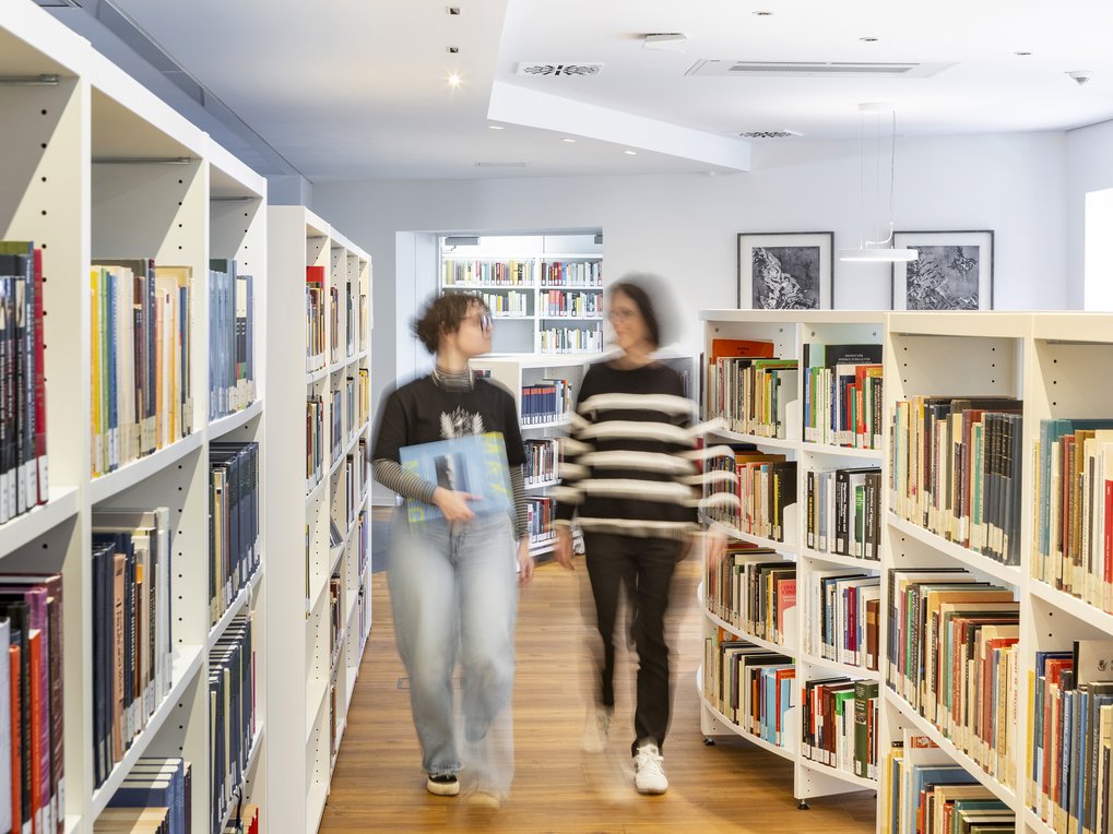 Zwei Personen bewegen sich durch eine moderne Bibliothek mit weißen Regalen, die mit vielen Büchern gefüllt sind, auf einem Holzfußboden.