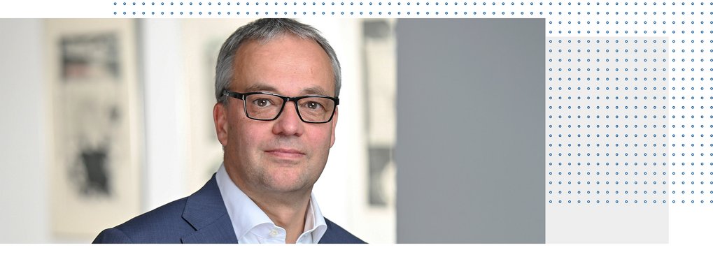 Ein Mann mit kurzen grauen Haaren und Brille, bekleidet mit einem blauen Anzug und einem weißen Hemd, posiert in einem Innenraum für die Kamera.
