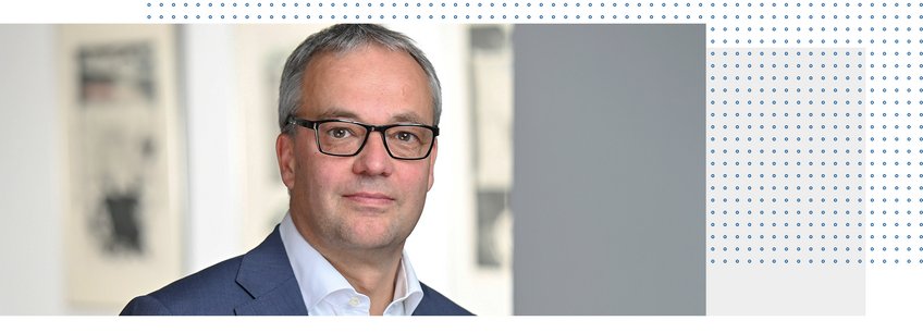 Prof. Dr. Jens Beckert Ein Mann mit kurzen grauen Haaren und Brille, bekleidet mit einem blauen Anzug und einem weißen Hemd, posiert in einem Innenraum für die Kamera.