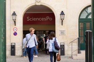 Eingang von SciencesPo, einer renommierten Universität, mit mehreren Personen, die den Campus betreten oder verlassen. Im Vordergrund sind Hinweisschilder und Laternen an der Wand sichtbar.
