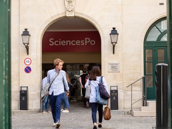 Foto von Marta Nascimento, Sciences Po (2019), Eingang Gebäude Rue de la Chaise Eingang von SciencesPo, einer renommierten Universität, mit mehreren Personen, die den Campus betreten oder verlassen. Im Vordergrund sind Hinweisschilder und Laternen an der Wand sichtbar.