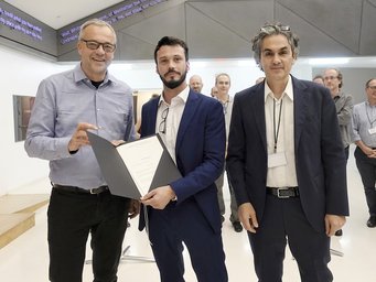 Jens Beckert, Felipe González López und Lucio Baccaro bei der Urkundenübergabe zur Gründung der Partnergruppe Drei Männer in Anzügen und mit Namensschildern stehen in einem modernen Innenraum; einer von ihnen hält eine geöffnete Mappe in der Hand, in der ein Dokument zu sehen ist.