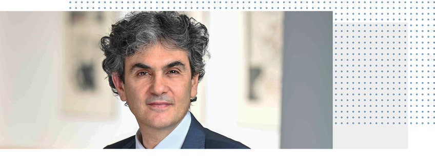 Prof. Dr. Lucio Baccaro Individual with curly hair, dressed in a dark suit against an abstract backdrop.