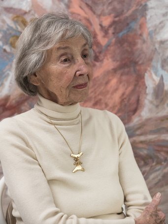 An elderly woman with gray hair, wearing a cream-colored turtleneck sweater and a gold necklace, is sitting against a colorful background.