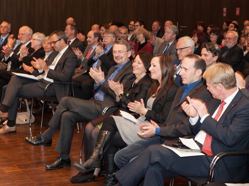 Eine Gruppe von Personen in Business-Anzügen und formeller Kleidung sitzt in einem Konferenzsaal und klatscht aufmerksam, was ihr Interesse an der Veranstaltung zeigt.