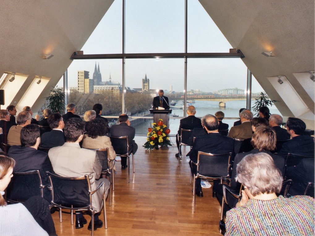 Ein Redner spricht vor einem sitzenden Publikum in einem modernen Veranstaltungsort mit großen dreieckigen Fenstern, die einen Blick auf den Kölner Dom und den Rhein bieten.