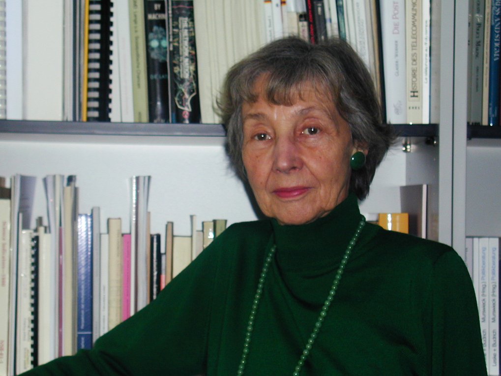 Person trägt einen grünen Rollkragenpullover und eine lange grüne Halskette. Im Hintergrund befindet sich ein Bücherregal mit verschiedenen Büchern.