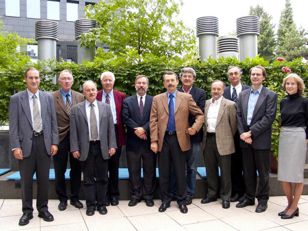 Zehn Personen, gekleidet in einer Mischung aus Anzügen und Business-Casual-Kleidung, stehen gemeinsam im Freien vor üppigem Grün und industriellen Lüftungsschächten.