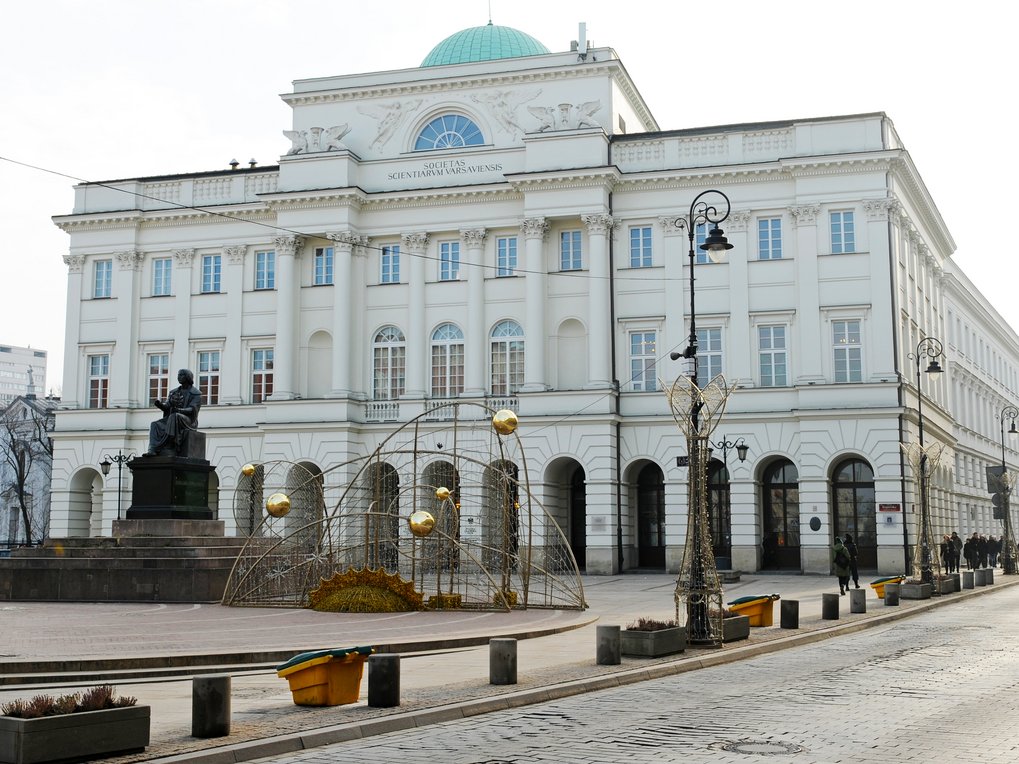 Neoklassizistisches Gebäude mit einer grünen Kuppel, einer sitzenden Statue auf einem Sockel und dekorativen Festbögen auf einem Stadtplatz, begleitet von Straßenlaternen und einer Kopfsteinpflasterstraße.