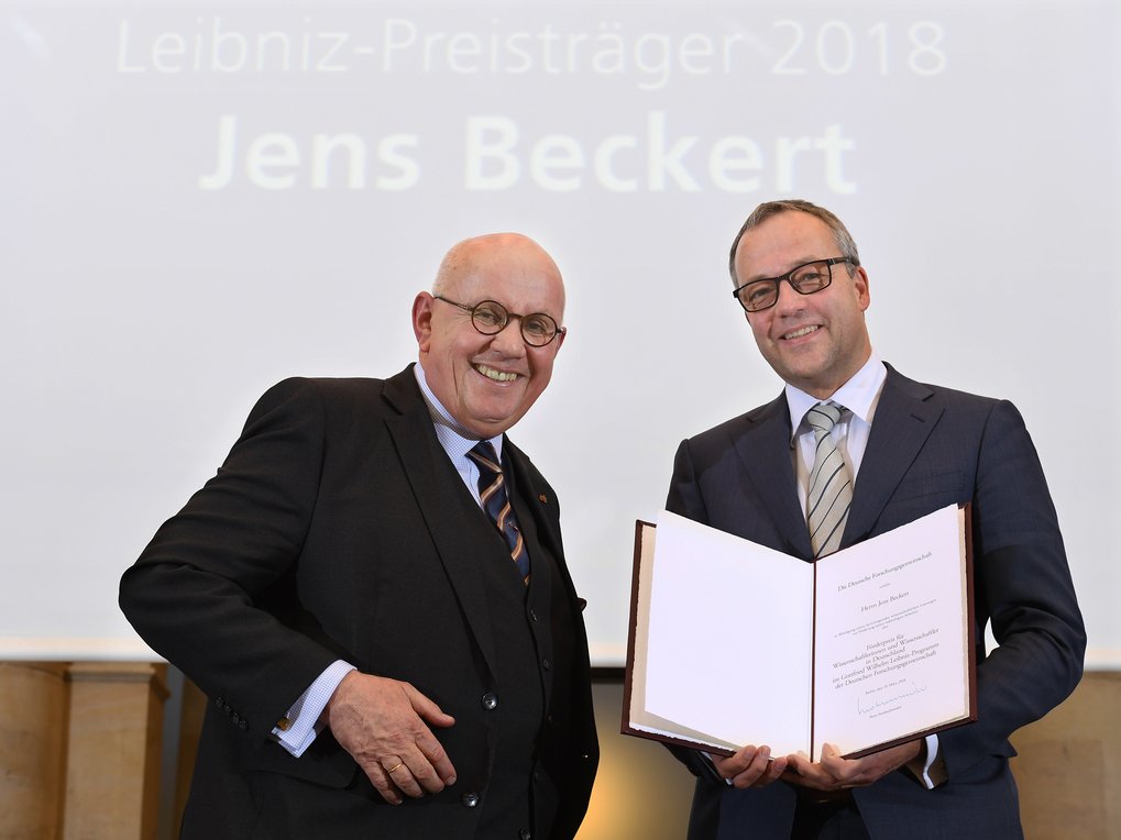 Zwei Männer in formeller Kleidung. Der Mann rechts (Jens Beckert) präsentiert eine geöffnete Urkunde. Im Hintergrund wird eine PowerPoint-Präsentation mit dem Text „Leibniz-Preisträger 2018: Jens Beckert” an die Wand projiziert.