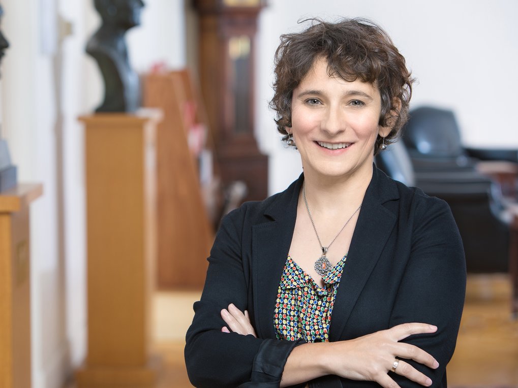 Frau (Marion Fourcade) mit lockigem Haar und schwarzem Blazer steht vor einer Büroeinrichtung. Im Hintergrund sind mehrere Büsten und ein Ledersessel.