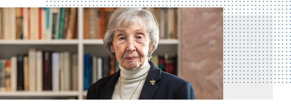 An elderly woman with gray hair is standing in front of a bookshelf filled with books. She is wearing a dark blazer and a light-colored turtleneck.