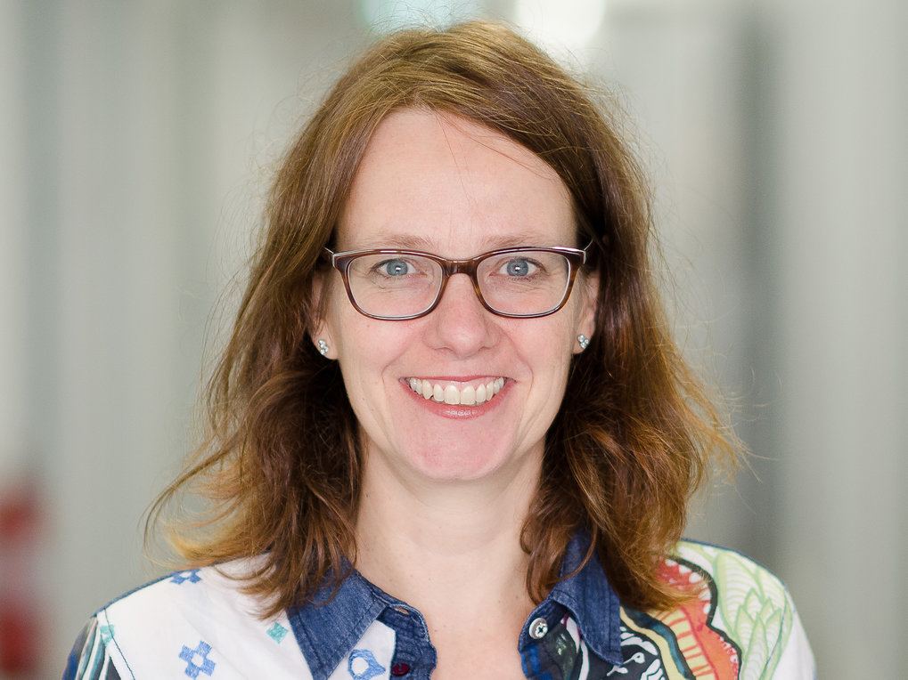 Eine Person mit braunen Haaren, vermutlich weiblichen Geschlechts, trägt ein Hemd mit floralen und geometrischen Mustern sowie silberne Ohrringe. Der Hintergrund ist verschwommen.