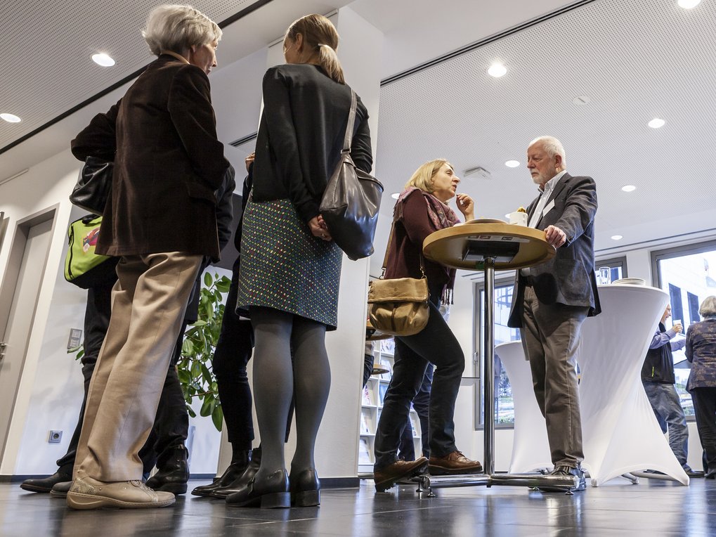 People at a corporate event indoors, interacting in small groups near a high table; some are holding bags, showcasing a professional setting.