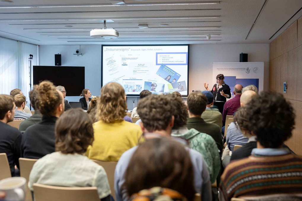 A group of people seated in a modern conference room, engaged with a presentation featuring slides on topics like discourse events, social media, and communication strategies.