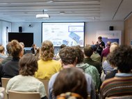 Eine Gruppe von Personen sitzt in einem modernen Konferenzraum und verfolgt eine Präsentation mit Folien zu Themen wie Diskursereignissen, sozialen Medien und Kommunikationsstrategien.