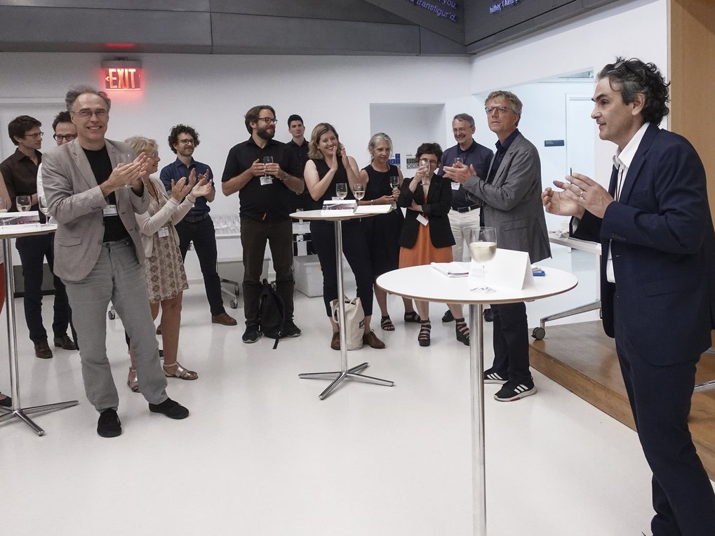 Attendees in business attire at a reception clapping, surrounded by high tables with wine glasses, in a contemporary setting.