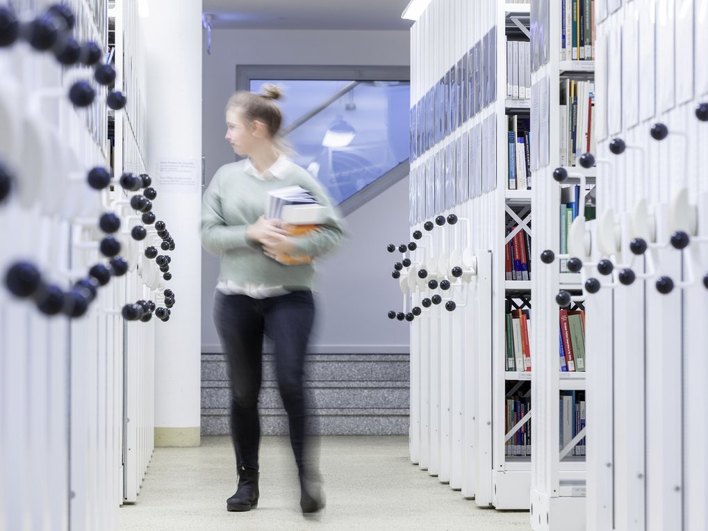 In a modern library, rows of white shelves house numerous books. An individual, obscured, carries documents, moving through the corridor.