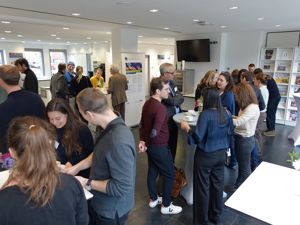Konferenzraum mit Zuhörern, Vortragende Person präsentiert Informationen auf einer Leinwand; Publikum sitzt aufmerksam.