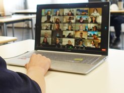 Ein Laptop, auf dessen Bildschirm eine Videokonferenz mit mehreren Teilnehmern zu sehen ist, von der Seite betrachtet, mit einer Hand auf der Tastatur.