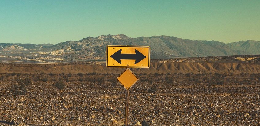 Die Zukunft in der Krise Ein gelbes Verkehrsschild mit zwei Pfeilen in entgegengesetzte Richtungen steht in einer kargen Wüstenlandschaft, im Hintergrund eine Bergkette unter klarem Himmel.