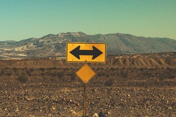 Ein gelbes Verkehrsschild mit zwei Pfeilen in entgegengesetzte Richtungen steht in einer kargen Wüstenlandschaft, im Hintergrund eine Bergkette unter klarem Himmel.