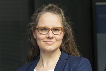 Eine Person mit langen Haaren trägt einen dunkelblauen Blazer über einem weißen, gepunkteten Oberteil, vor einem dunklen Hintergrund.