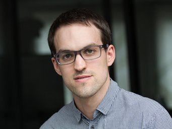 Person mit kurzen dunklen Haaren und einem weiß-blauen karierten Hemd vor einem dunklen, unscharfen Hintergrund.