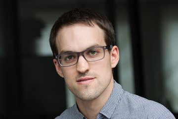 Person mit kurzen dunklen Haaren und einem weiß-blauen karierten Hemd vor einem dunklen, unscharfen Hintergrund.