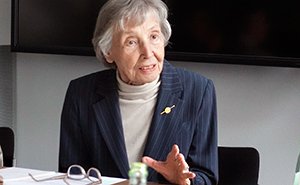 Eine Person in einem gestreiften Anzug mit Brosche sitzt an einem Tisch und gestikuliert, auf dem Tisch liegen Dokumente und eine Brille.