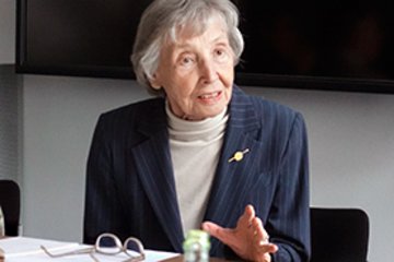 Eine Person in einem gestreiften Anzug mit Brosche sitzt an einem Tisch und gestikuliert, auf dem Tisch liegen Dokumente und eine Brille.