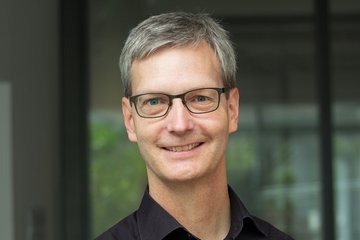 Mann mit grauem Haar und schwarzem Hemd, im Hintergrund eine Glasfassade mit grünen Pflanzen.