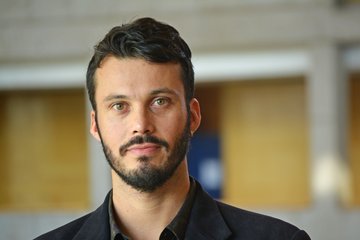 Ein Mann mit dunklen lockigen Haaren und einem schwarzen Anzug steht vor einem verschwommenen Hintergrund in einem Gebäude.