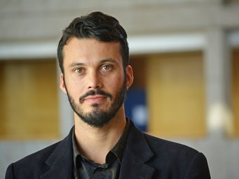 Ein Mann mit dunklen lockigen Haaren und einem schwarzen Anzug steht vor einem verschwommenen Hintergrund in einem Gebäude.