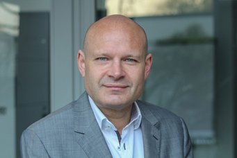 Eine Person in einem grauen Anzug mit offenem weißen Hemd steht in einem modernen Innenraum mit einer Glaswand im Hintergrund.