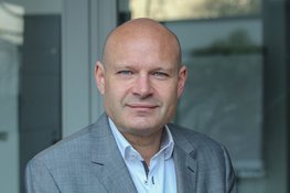 Eine Person in einem grauen Anzug mit offenem weißen Hemd steht in einem modernen Innenraum mit einer Glaswand im Hintergrund.