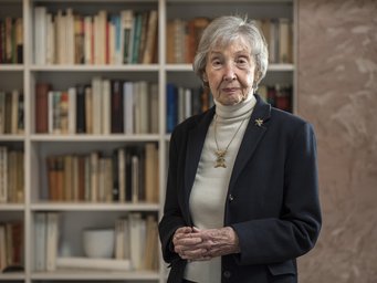 Eine ältere Person, gekleidet in einen dunklen Blazer und einen hellen Rollkragenpullover, steht vor einem großen Bücherregal, das mit verschiedenen Büchern gefüllt ist. Im Hintergrund sind die Bücher in unterschiedlichen Größen und Farben zu sehen. Die Person trägt eine goldene Halskette und eine Brosche.