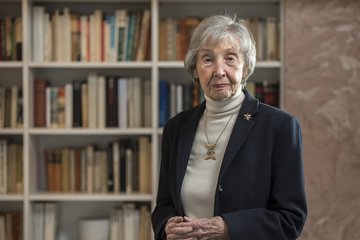 Eine ältere Person, gekleidet in einen dunklen Blazer und einen hellen Rollkragenpullover, steht vor einem großen Bücherregal, das mit verschiedenen Büchern gefüllt ist. Im Hintergrund sind die Bücher in unterschiedlichen Größen und Farben zu sehen. Die Person trägt eine goldene Halskette und eine Brosche.