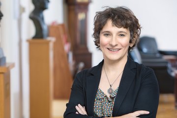 Frau im Büro, trägt schwarzen Blazer und gemustertes Oberteil, steht mit verschränkten Armen vor Skulpturen und Büromöbeln.