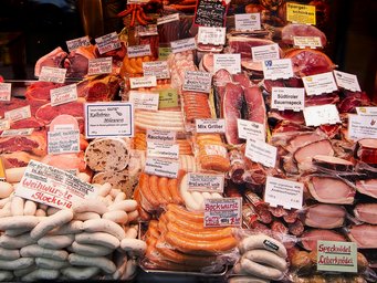 Eine Auslage mit verschiedenen Fleischwaren wie Würsten, Schinken und Speck, versehen mit Preisschildern und Produktbeschreibungen.
