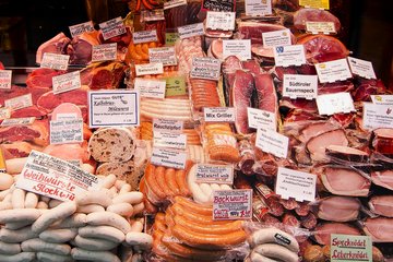 Eine Auslage mit verschiedenen Fleischwaren wie Würsten, Schinken und Speck, versehen mit Preisschildern und Produktbeschreibungen.