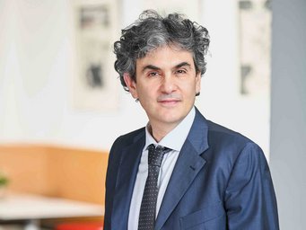 Mann mit grauem Haar, formellem Anzug und gepunkteter Krawatte in einem hellen, modernen Büro mit abstrakter Kunst.