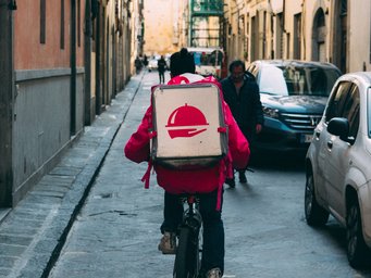 Ein Fahrradkurier in roter Jacke radelt durch eine belebte, enge Stadtstraße, während er eine weiße Lieferbox mit einem roten Symbol auf dem Rücken trägt. Autos parken am Straßenrand.