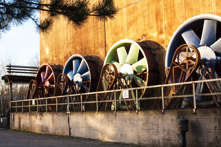 Deindustrialisierung als Fakt und Fiktion Eine Reihe von vier großen Ventilatoren in verschiedenen Farben, montiert an einer Betonwand, umgeben von einem Geländer. Ein Baumast im Vordergrund.
