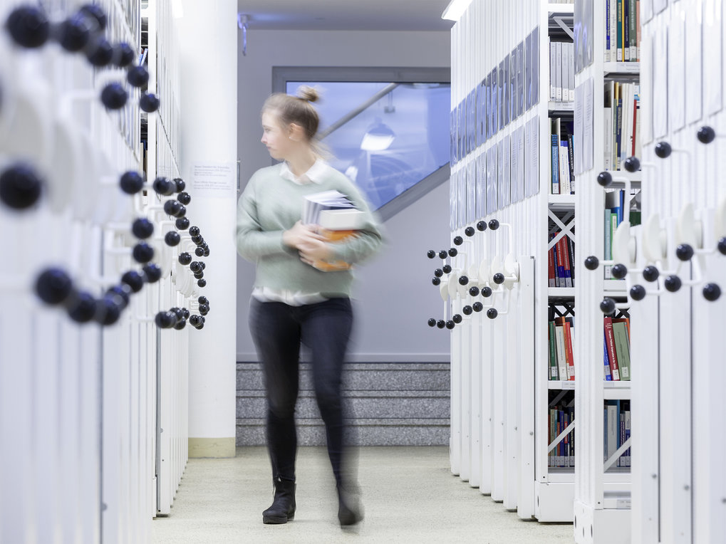 Eine Person trägt mehrere Bücher durch einen schmalen Gang, flankiert von hohen Bücherregalen, in einem gut beleuchteten Archivraum.