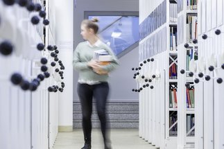 Eine Person trägt mehrere Bücher durch einen schmalen Gang, flankiert von hohen Bücherregalen, in einem gut beleuchteten Archivraum.