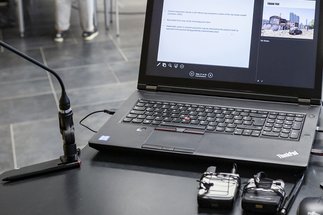 Ein Laptop mit geöffnetem Dokument und Bild auf einem Tisch, flankiert von Mikrofonen, im Hintergrund Stühle und stehende Personen.