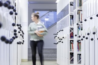 An individual moves through a library corridor, holding multiple books, surrounded by mobile shelving units with visible book spines.