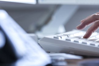 A close-up image shows a person's hand actively typing on a white keyboard, while a phone is blurred in the foreground, suggesting an office environment.