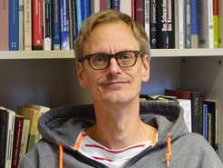 Vor einem Bücherregal mit politischer Literatur sitzt eine Person in einem grauen Kapuzenpulli mit orangefarbenen Kordeln.